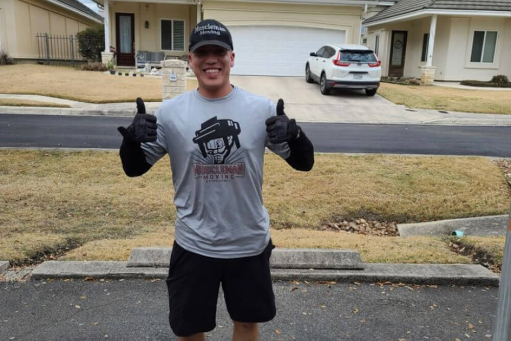 Photo of a man wearing black shorts, a grey Muscle Man Moving t-shirt, black gloves, and a black Muscle Man Moving cap giving a thumbs up with both hands and smiling.