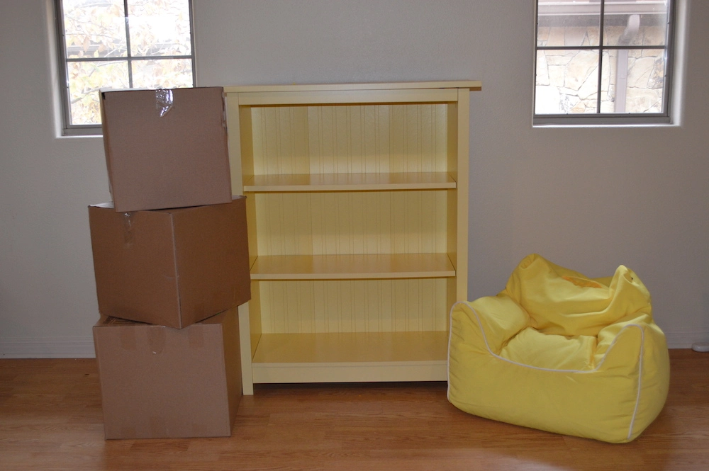 A three tiered yellow shelf, yellow bean bag, and three cardboard boxes on the floor of an empty home.